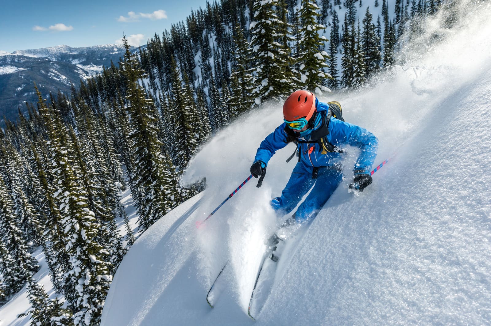 A skier in a blue jacket throwing up powder on a steep slope among pines
